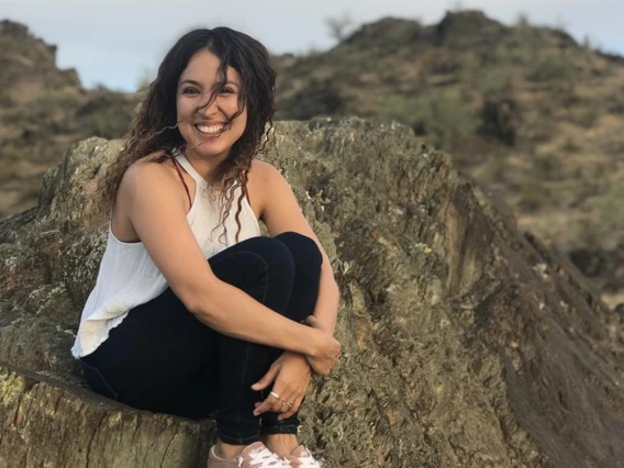 Alba Porras smiling and sitting on a rocky ledge outdoors, with desert hills and cloudy sky in the background.