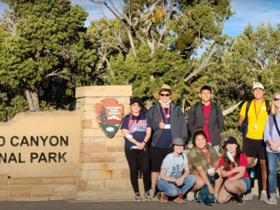 A group of students poses in front of a "Grand Canyon National Park" sign with trees in the background, wearing visitor lanyards.