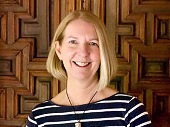 Barbara Mills smiling while standing in front of an ornate wooden panel wall, wearing a navy and white striped top and a pendant necklace.