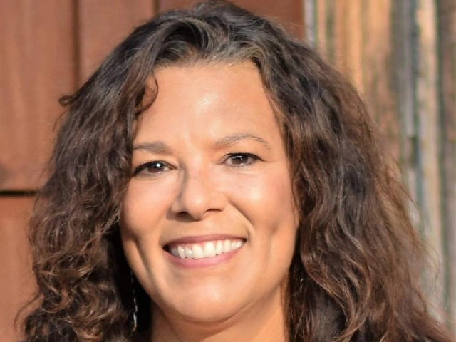 Corey Knox smiling in front of a sunlit wooden wall, wearing a black top with wavy shoulder-length hair.