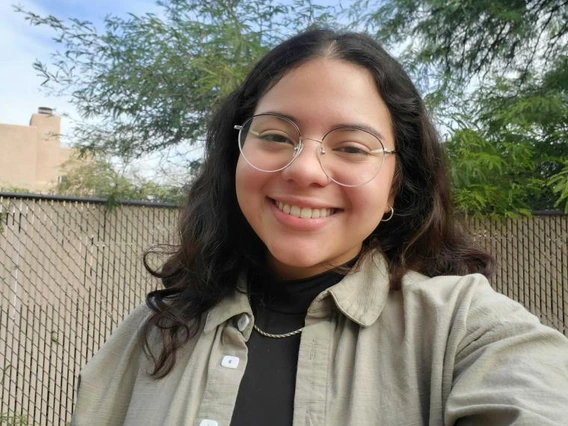 Karla Paredes smiling outdoors in front of a fence and trees, wearing round glasses, a tan overshirt, and a black top