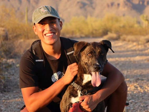 Omar Solis smiling while kneeling beside a happy dog with a red leash on a desert trail, with mountains and cactus in the background at sunset.