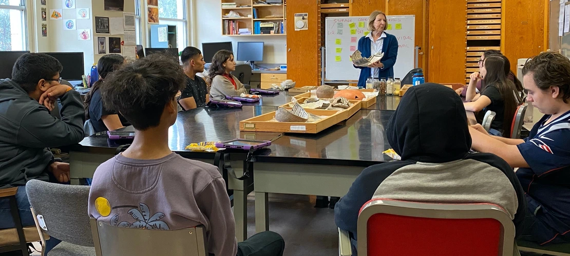 A group of students sits around a large table listening to an instructor holding pottery artifacts in a classroom filled with boxes and display trays.