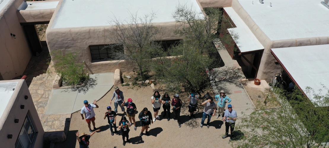Aerial view of students and instructors waving at a drone in a desert courtyard between adobe-style buildings.