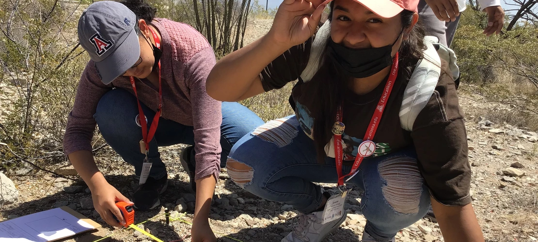 Two students crouch in a desert landscape using a measuring tape during a field activity, wearing hats and red lanyards.