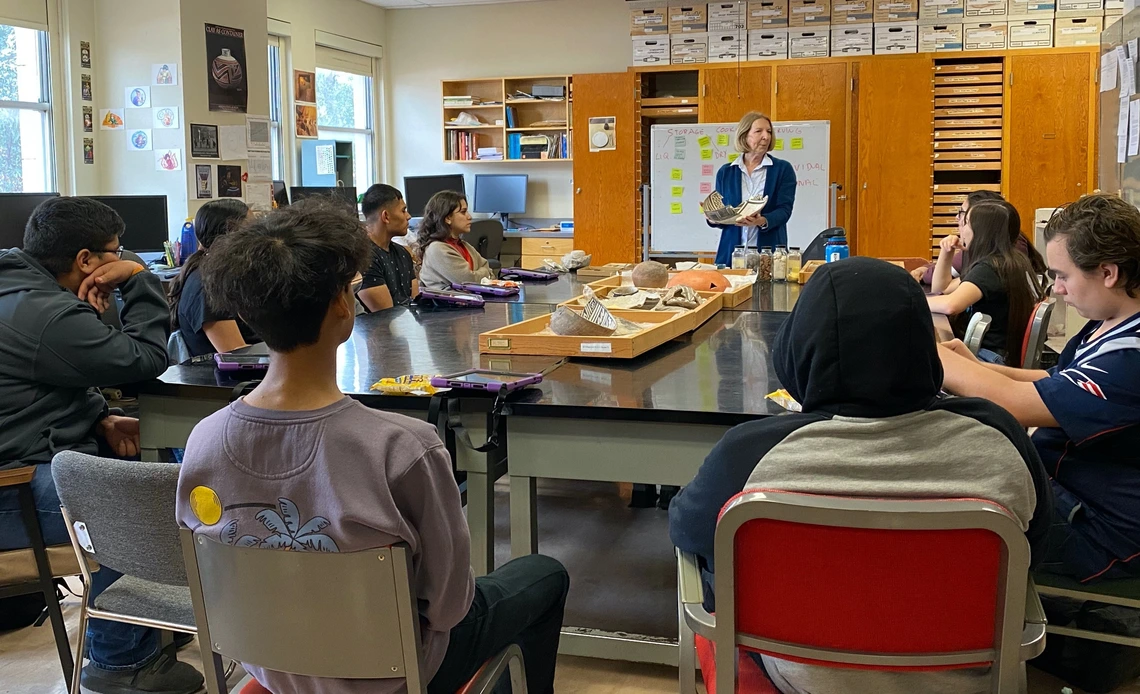 A group of students sits around a large table listening to an instructor holding pottery artifacts in a classroom filled with boxes and display trays.