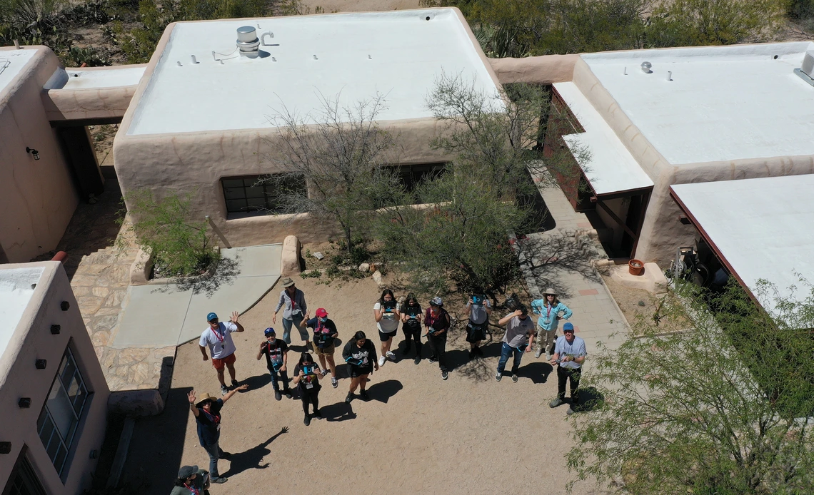 Aerial view of students and instructors waving at a drone in a desert courtyard between adobe-style buildings.