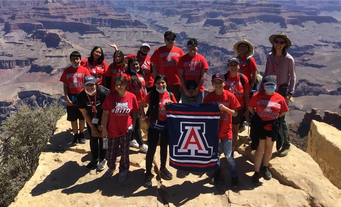 A group of students and instructors wearing red University of Arizona shirts poses with a UA flag at the edge of the Grand Canyon.