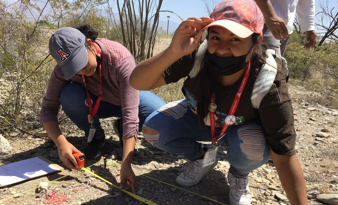 Two students crouch in a desert landscape using a measuring tape during a field activity, wearing hats and red lanyards.