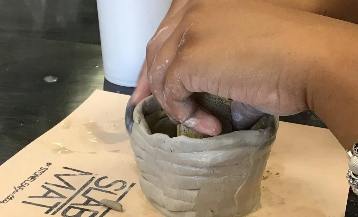 Close-up of a student's hands shaping a small coiled clay pot on a work mat during a pottery-making activity.