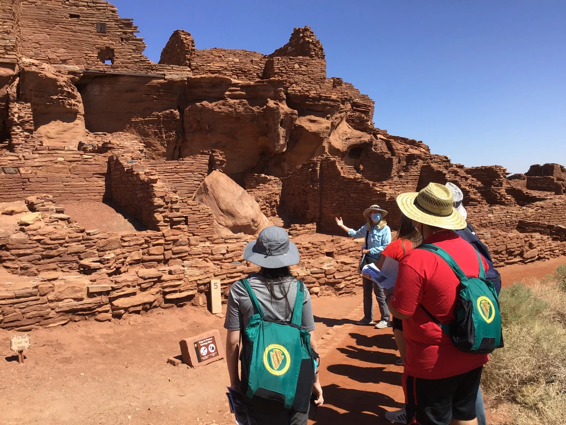 A group of students with green backpacks listens to a guide at ancient stone ruins under a clear blue sky, likely at a Southwest archaeological site.