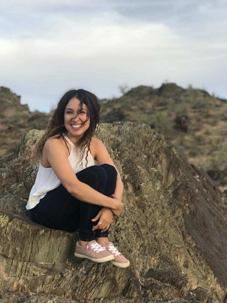 Alba Porras smiling and sitting on a rocky ledge outdoors, with desert hills and cloudy sky in the background.