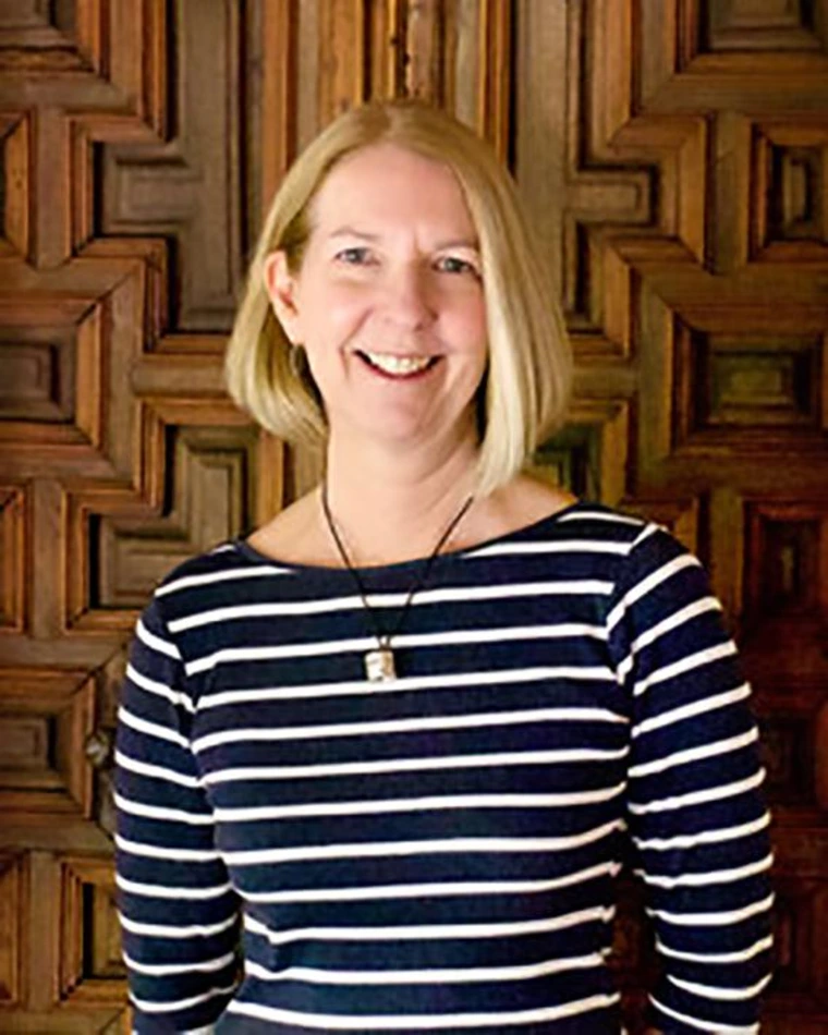 Barbara Mills smiling while standing in front of an ornate wooden panel wall, wearing a navy and white striped top and a pendant necklace.
