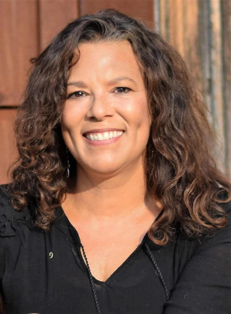 Corey Knox smiling in front of a sunlit wooden wall, wearing a black top with wavy shoulder-length hair.