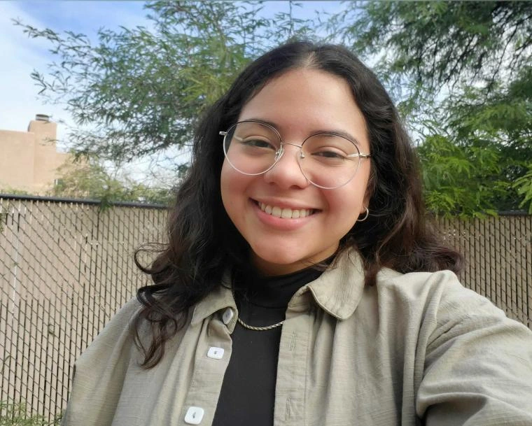 Karla Paredes smiling outdoors in front of a fence and trees, wearing round glasses, a tan overshirt, and a black top