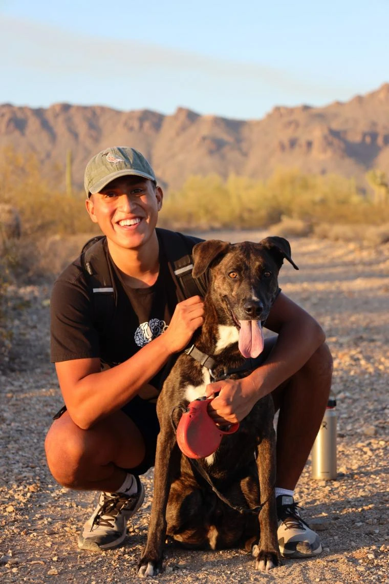 Omar Solis smiling while kneeling beside a happy dog with a red leash on a desert trail, with mountains and cactus in the background at sunset.