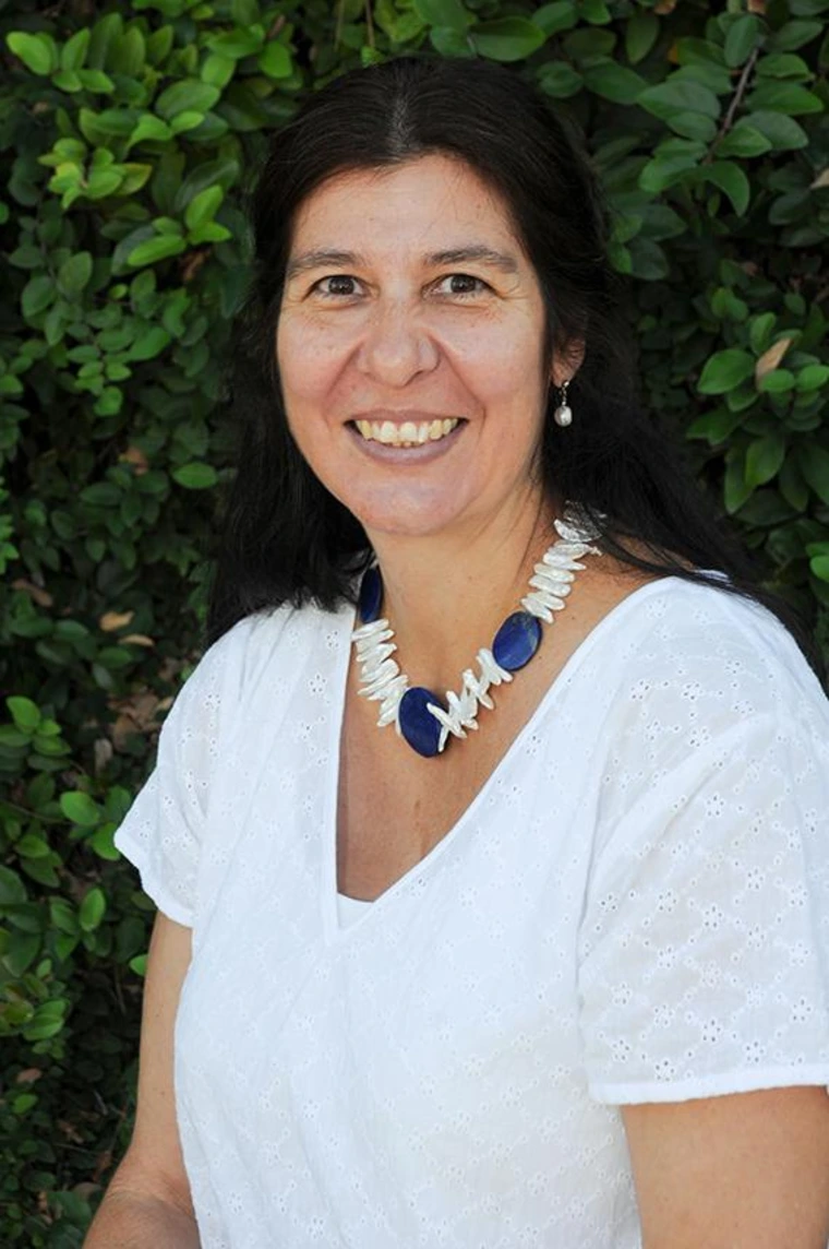 Sara Chavarria smiling outdoors in front of leafy green foliage, wearing a white blouse and a blue and white beaded necklace
